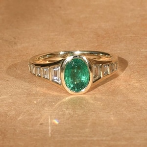 Puede incluir: Un anillo de oro con una piedra central de esmeralda ovalada, flanqueada por diamantes talla baguette. El anillo tiene una banda de oro pulido y un diseño clásico y elegante. La esmeralda es de color verde vibrante.