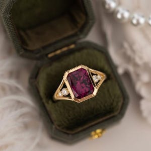 Könnte beinhalten: Ein goldener Ring mit einem großen, rechteckigen, tiefroten Edelstein in der Mitte. Der Ring ist in einem goldenen Band mit kleinen, klaren Akzentsteinen auf beiden Seiten des Hauptsteins gefasst. Der Ring wird in einer grünen Samt-Ringbox präsentiert.