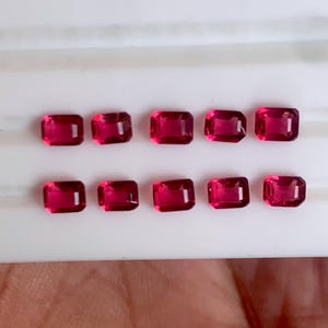 May include: Ten rectangular, faceted, deep red gemstones are arranged in two rows. The gems have a vibrant, saturated color and a polished appearance. The gems are set against a white background.