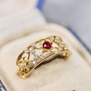 Könnte beinhalten: Ein goldfarbener Ring mit offenem Filigran-Design. Der Ring hat einen zentralen, runden, roten Edelstein und ist mit kleinen, klaren Edelsteinen verziert. Der Ring wird in einer hellen Schmuckschatulle präsentiert.