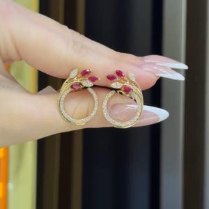 Könnte beinhalten: Goldfarbene Ohrringe mit kreisförmigem Design, verziert mit kleinen roten Edelsteinen und klaren Kristallen. Die Ohrringe haben ein astähnliches Motiv, wobei die roten Steine an Beeren erinnern. Die Kristalle sind entlang des kreisförmigen Rahmens gesetzt.