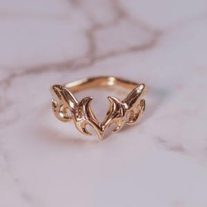 Puede incluir: Un anillo dorado con un diseño decorativo. El anillo presenta una forma central estilizada de llama o corazón, flanqueada por elementos curvos ornamentados. El anillo está sobre un fondo de mármol.