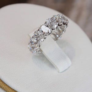 Könnte beinhalten: Ein silberner Ring mit einer Reihe von oval geschliffenen Diamanten. Die Diamanten funkeln hell und reflektieren das Licht. Der Ring wird auf einer weißen Oberfläche präsentiert, die sein elegantes Design hervorhebt. Ein Schmuckstück.