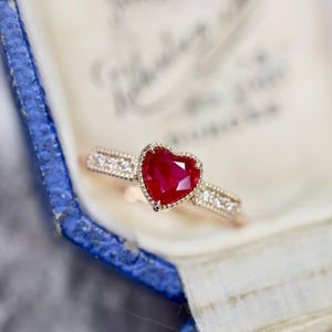 Op de afbeelding: Een gouden ring met een hartvormige, gefacetteerde rode edelsteen. De ringband is versierd met kleine, heldere edelstenen. De ring wordt gepresenteerd in een blauwe doos met een wit interieur.