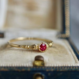 Könnte beinhalten: Ein goldener Ring mit einem kleinen, runden, roten Edelstein. Der Ring ist in einer dekorativen Goldeinfassung gefasst und wird in einer blauen Schmuckschatulle mit Samt präsentiert. Das Band des Rings hat ein filigranes Design.