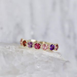 Puede incluir: Un anillo de oro con una fila de piedras preciosas de colores en tonos rojos, rosas y morados. Las piedras preciosas redondas están engastadas juntas, creando un diseño vibrante. El anillo se muestra sobre una superficie blanca texturizada.