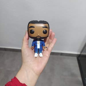 Könnte beinhalten: Eine Funko Pop Vinylfigur einer Person mit dunkler Haut, die eine blaue Jacke mit weißen Akzenten, ein weißes Hemd und weiße Schuhe trägt. Die Figur hat eine goldene Kette und das Wort "Faith" auf der Seite des Kopfes.