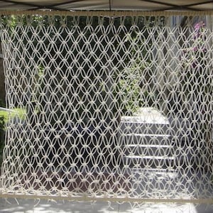 Puede incluir: Colgante de pared de macramé color crema con un patrón geométrico. El diseño tejido presenta formas ovaladas interconectadas, creando una pantalla calada texturizada. El macramé está suspendido de una barra de madera, adecuado para la decoración interior o exterior.