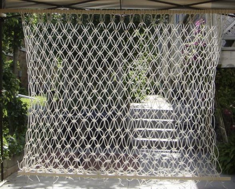 Op de afbeelding: Cr&egrave;mekleurige macram&eacute; wandhanger met een geometrisch patroon. Het geweven ontwerp heeft onderling verbonden ovale vormen, waardoor een gestructureerd, open scherm ontstaat. De macram&eacute; is opgehangen aan een houten staaf, geschikt voor binnen- of buitendecoratie.
