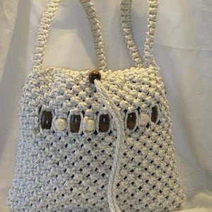 May include: Off-white macrame handbag with two handles and a drawstring closure. The bag features a decorative row of wooden and off-white beads. The macrame weave creates a textured, open pattern. A stylish, handmade accessory.