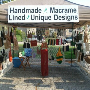 Op de afbeelding: Een marktkraam met handgemaakte macram&eacute; tassen in diverse stijlen en kleuren. Het bord leest "Handmade Macrame Lined Unique Designs". De tassen omvatten schoudertassen, draagtassen en modellen met franjes, die een reeks texturen en patronen laten zien.