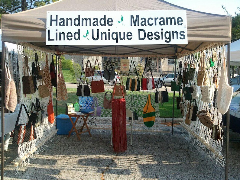 Op de afbeelding: Een marktkraam met handgemaakte macram&eacute; tassen in diverse stijlen en kleuren. Het bord leest "Handmade Macrame Lined Unique Designs". De tassen omvatten schoudertassen, draagtassen en modellen met franjes, die een reeks texturen en patronen laten zien.