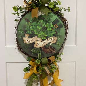 May include: Round St. Patrick's Day wreath with a green and brown color scheme. The wreath features a central image of shamrocks, a leprechaun hat, and a banner that reads "Luck o' the Irish." It is decorated with green and gold ribbons and faux greenery.