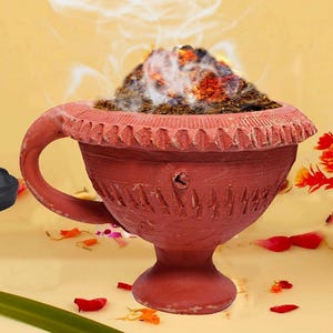 May include: A terracotta incense burner with a handle, filled with burning incense, emitting smoke. The burner has a decorative rim and etched patterns. The text "GAU VANSH" is visible. A small black statue and a vase of orange flowers are in the background.