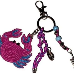 May include: A keychain featuring a pink beaded crab charm with blue accents. Additional charms include a seashell, a turtle, and a beaded strand with light blue beads. The keychain has a silver clasp and a key ring.