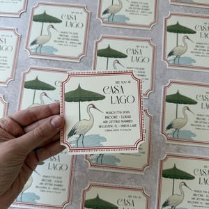 May include: Wedding invitation cards with a Casa Lago theme. Each card features a crane illustration under a green parasol. The cards have a red border and include event details: date, names, and location.
