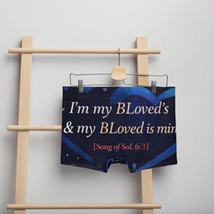 May include: Dark blue boxer briefs with the text "I'm my BLoved's & my BLoved is min [Song of Sol. 6:3]" in white. The underwear is hanging on a wooden hanger and a wooden ladder.