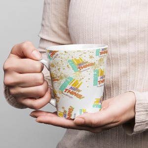 Może przedstawiać: Biały ceramiczny kubek z powtarzającym się wzorem słów "NEW THING!" w kolorze żółtym, czerwonym i niebieskim, z efektem rozprysku. Kubek ma biały uchwyt i jest trzymany w dłoniach osoby.