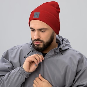 May include: A red beanie with a gray square patch featuring a circular design. The beanie is ribbed and appears to be made of a soft material. The hat is worn by a person wearing a gray jacket.