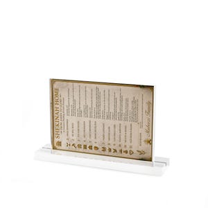May include: A framed print with the text "SHEKINAH HOME" and other text, displayed on a white stand. The print has a beige background with gold-colored borders and text. The text includes "Mubeezi Family".