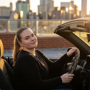 May include: A black steering wheel cover featuring a repeating geometric pattern of tan and dark brown triangles. The cover is fitted onto a circular steering wheel inside a vehicle, providing a textured grip surface for the driver to hold while operating.