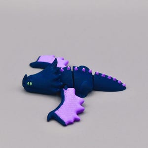 Puede incluir: Figura de drag&oacute;n azul oscuro con detalles morados, incluyendo alas y p&uacute;as. El drag&oacute;n tiene ojos verdes y est&aacute; segmentado. Probablemente un juguete o un art&iacute;culo decorativo.