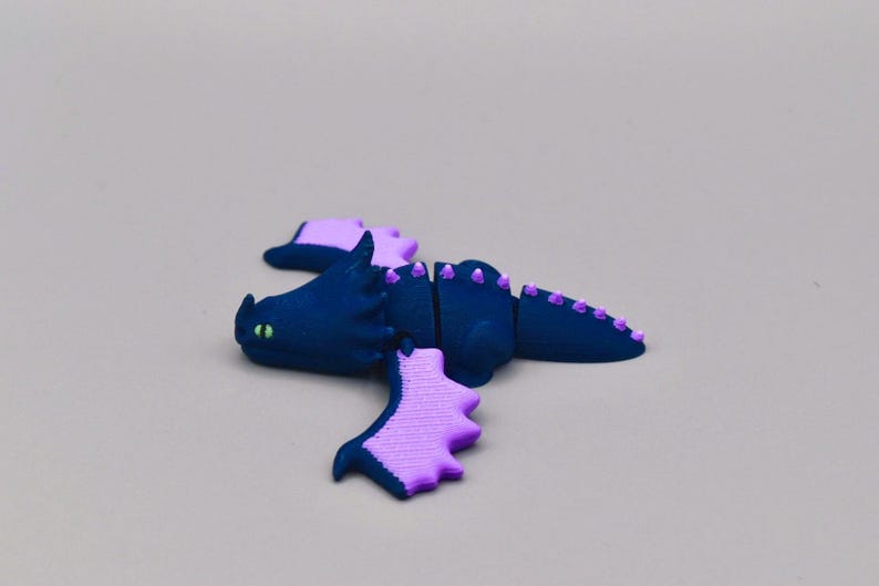 Puede incluir: Figura de drag&oacute;n azul oscuro con detalles morados, incluyendo alas y p&uacute;as. El drag&oacute;n tiene ojos verdes y est&aacute; segmentado. Probablemente un juguete o un art&iacute;culo decorativo.