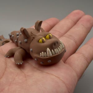 May include: A brown, 3D-printed creature figurine with yellow eyes and white teeth. The figurine has small gray bumps along its body and is held in an open hand. The creature has small legs and ears.