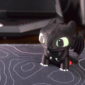 以下が含まれることがあります： 大きな緑色の目と小さな翼を持つ黒いドラゴンの置物。 ドラゴンは足に白いアクセントがあり、赤い尾びれがあります。 白黒の模様の表面に座っています。