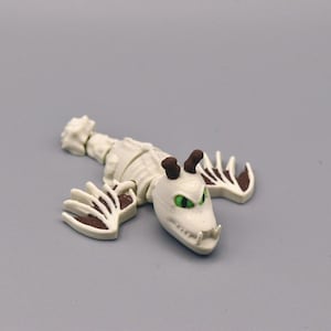 Puede incluir: Figura de drag&oacute;n esquel&eacute;tico blanco con detalles marrones en garras y cuernos. El drag&oacute;n tiene ojos verdes y un dise&ntilde;o detallado, sobre una superficie gris.