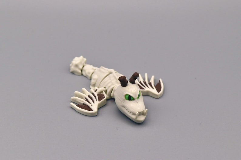 Puede incluir: Figura de drag&oacute;n esquel&eacute;tico blanco con detalles marrones en garras y cuernos. El drag&oacute;n tiene ojos verdes y un dise&ntilde;o detallado, sobre una superficie gris.