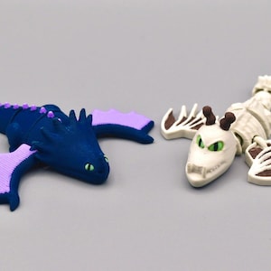 Könnte beinhalten: Zwei Drachenfiguren. Eine ist dunkelblau mit lila Flügeln und Akzenten, die andere ist ein weißes Skelettdrachen mit braunen und weißen Akzenten. Beide haben grüne Augen und befinden sich auf einer grauen Oberfläche.