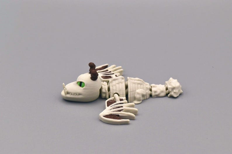 Puede incluir: Una figura de esqueleto de drag&oacute;n segmentada, blanquecina, con detalles marrones. El drag&oacute;n tiene ojos verdes y peque&ntilde;os cuernos marrones. La figura est&aacute; sobre una superficie gris.
