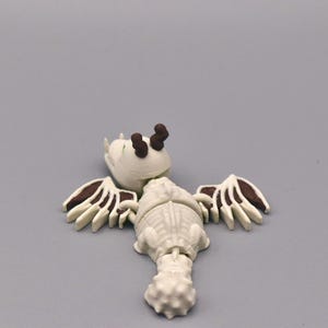Puede incluir: Una figura blanca y marr&oacute;n de una criatura con alas, una cabeza con dos cuernos marrones y un cuerpo segmentado. Las alas tienen detalles marrones. La figura est&aacute; sobre una superficie gris.