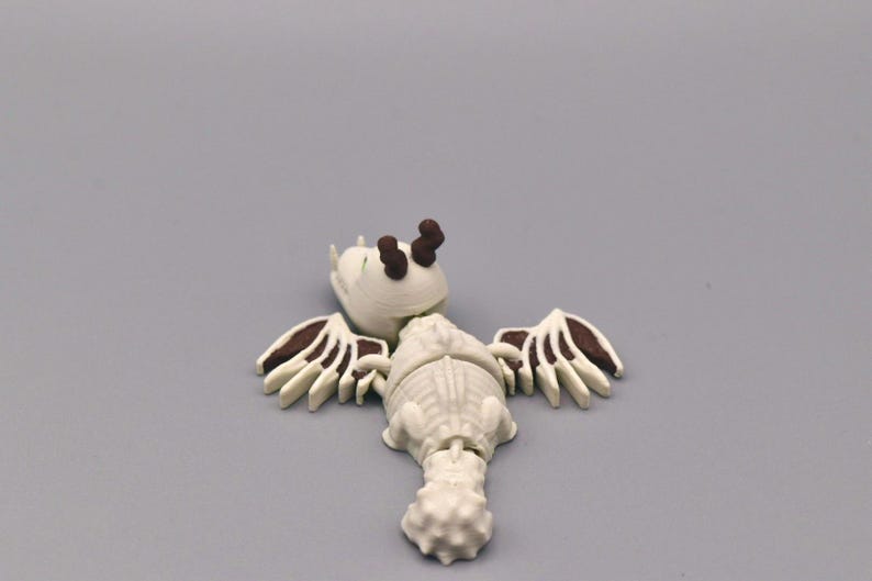 Puede incluir: Una figura blanca y marr&oacute;n de una criatura con alas, una cabeza con dos cuernos marrones y un cuerpo segmentado. Las alas tienen detalles marrones. La figura est&aacute; sobre una superficie gris.