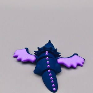 Puede incluir: Figura de drag&oacute;n azul oscuro con alas y detalles en lavanda. El drag&oacute;n tiene una cabeza y cola puntiagudas, con una l&iacute;nea de puntos lavanda en la espalda. Las alas est&aacute;n extendidas, sobre un fondo gris.