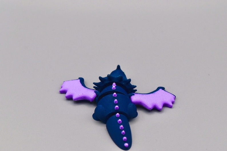 Puede incluir: Figura de drag&oacute;n azul oscuro con alas y detalles en lavanda. El drag&oacute;n tiene una cabeza y cola puntiagudas, con una l&iacute;nea de puntos lavanda en la espalda. Las alas est&aacute;n extendidas, sobre un fondo gris.