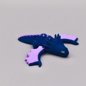 Puede incluir: Una figura de drag&oacute;n azul marino con alas y detalles en lavanda. El drag&oacute;n tiene ojos verdes y una fila de p&uacute;as lavanda en la espalda. Las alas tienen un dise&ntilde;o festoneado. La figura est&aacute; sobre una superficie gris claro.