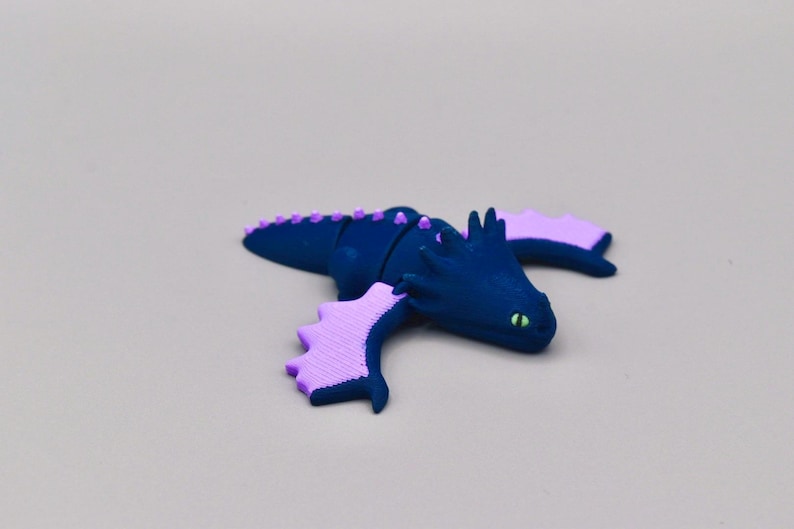 Puede incluir: Una figura de drag&oacute;n azul marino con alas y detalles en lavanda. El drag&oacute;n tiene ojos verdes y una fila de p&uacute;as lavanda en la espalda. Las alas tienen un dise&ntilde;o festoneado. La figura est&aacute; sobre una superficie gris claro.