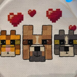 May include: Cross-stitch embroidery featuring three pixelated dog faces in shades of orange, brown, and gray, with red heart accents. The craft is on white fabric.