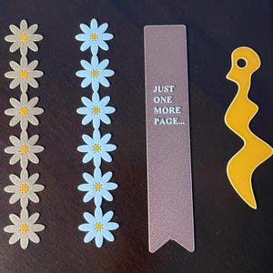 May include: An assortment of paper crafts including two daisy chains, a bookmark, and a yellow abstract shape. The daisy chains feature alternating colors of light gray and light blue flowers with yellow centers. The bookmark reads "JUST ONE MORE PAGE..."