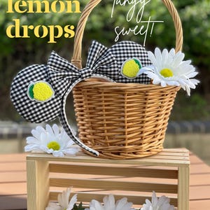 May include: A woven basket with a black and white checkered bow and matching ears, each adorned with a lemon applique. The text "Lemon Drops" and "Tangy sweet!" are visible. White daisies and a wooden crate complete the display.