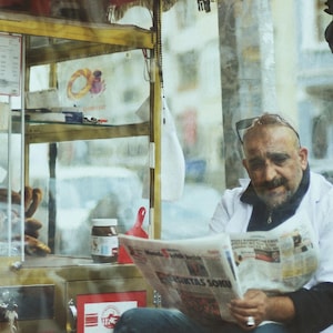 May include: A man reading a newspaper in front of a food cart. The cart has glass display cases with baked goods and a jar of hazelnut spread. The man is wearing a white coat and has sunglasses on his head.