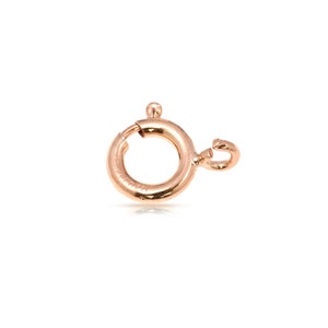 Puede incluir: Un cierre de anillo de resorte de oro rosa, un componente de joyería que se utiliza para asegurar collares y pulseras. El cierre es circular con una pequeña palanca para abrir y cerrar. El metal tiene un acabado brillante sobre un fondo blanco.
