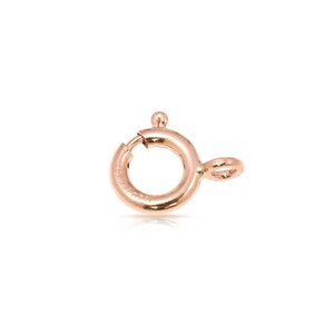 Puede incluir: Un cierre de anillo de resorte de oro rosa para la elaboración de joyas. El cierre es un círculo con un pequeño bucle en un lado y un cierre de resorte en el otro.