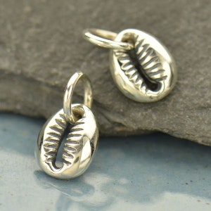 May include: Two silver-colored cowrie shell charms with a textured surface and a loop for hanging. The charms are displayed on a gray stone surface. These charms are suitable for jewelry making.