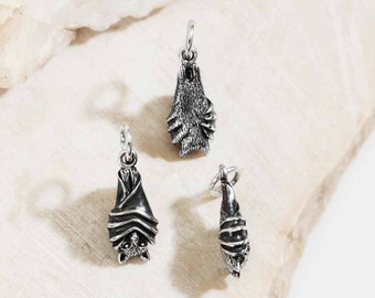 May include: Three silver bat charms with detailed designs. One bat is upright, another is hanging upside down, and the third is curled up. Each charm has a small loop for attaching to a necklace or bracelet. The charms are displayed on a light-colored, textured surface.