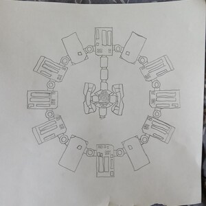 May include: A hand-drawn sketch of a circular space station design. The central hub connects to a ring of rectangular modules, each with detailed panels and circular attachments. The drawing is on a white sheet of paper.