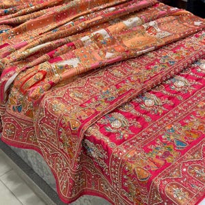 May include: A display of intricately embroidered textiles in shades of red, orange, and pink. The fabrics feature detailed patterns, embellished with beads and sequins. The textiles are folded and layered, showcasing the rich colors and textures of the designs.