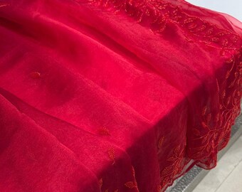 May include: A vibrant red fabric with intricate embroidered leaf patterns along the edges. The sheer material drapes over a surface, showcasing the delicate details of the embroidery. The fabric's rich color and design suggest it could be used for clothing or decorative purposes.